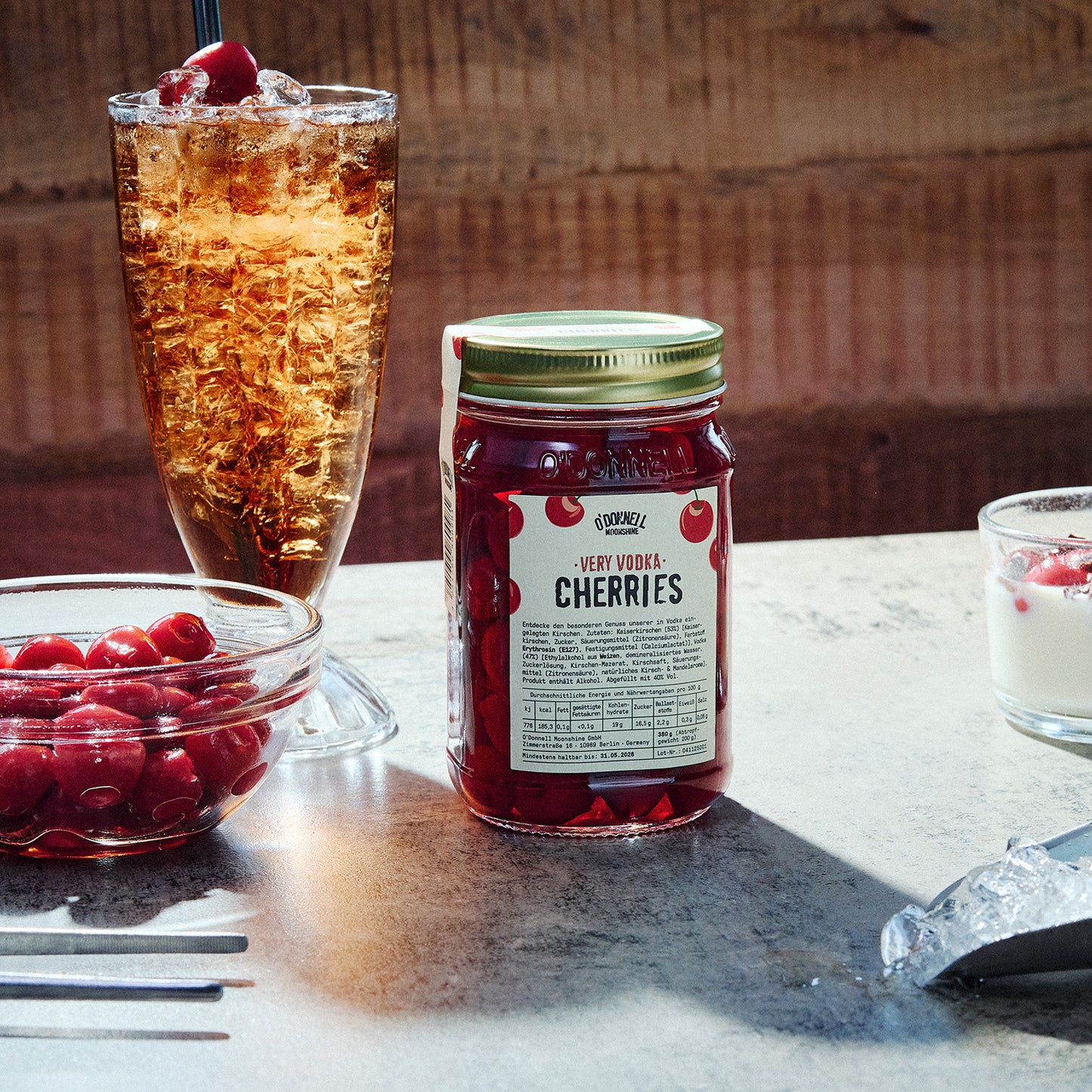 Jar of Very Vodka Cherries against a wooden background next to an iced drink and a bowl of cherries