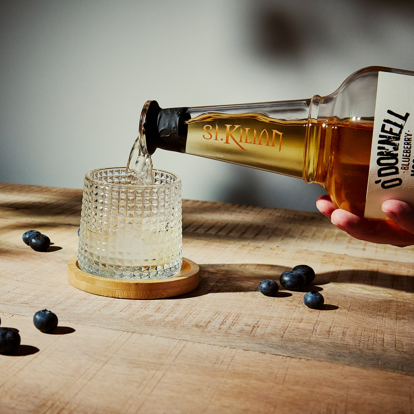 Bottle of St. Kilian blueberry liqueur being poured into a glass on a wooden surface with blueberries around.