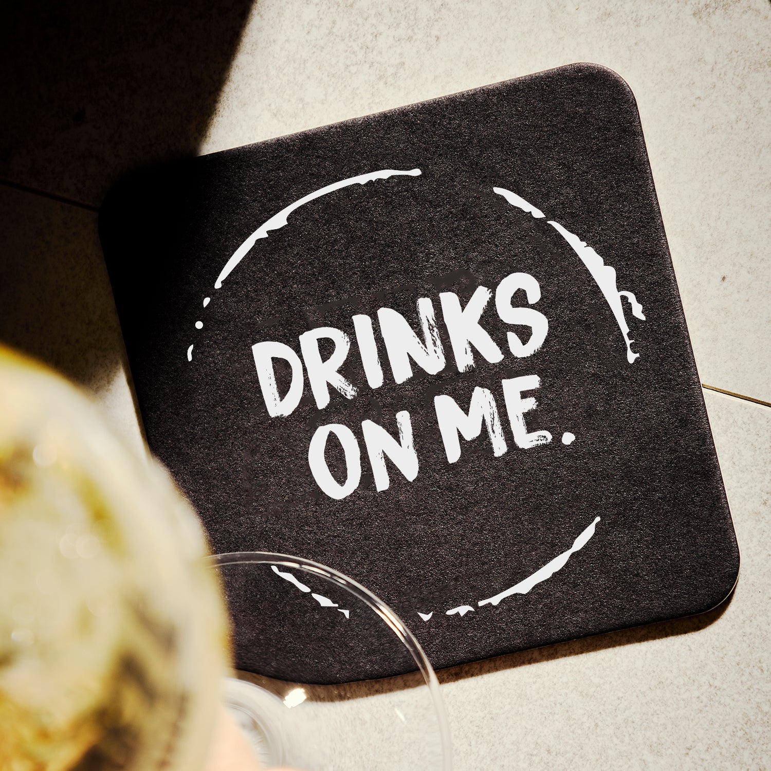 Black coaster with 'DRINKS ON ME.' text on a light surface