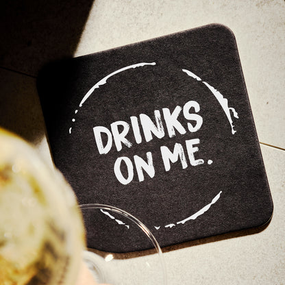 Black coaster with 'DRINKS ON ME.' text on a light surface