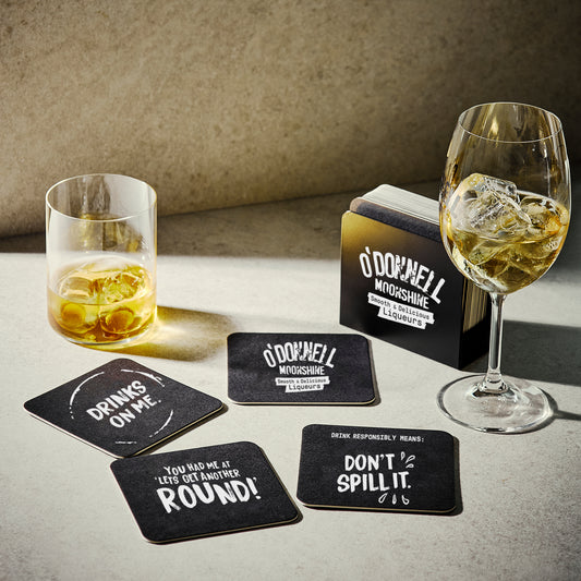 Set of black coasters with humorous sayings next to glasses of alcohol on a table.