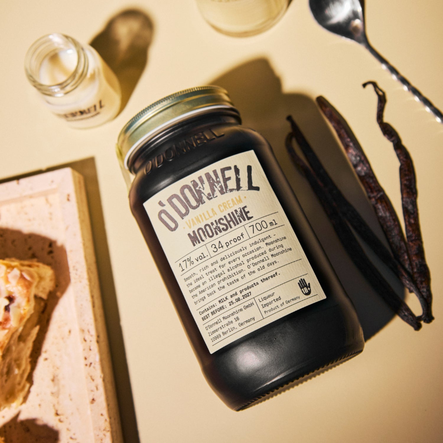 Vanilla Cream Moonshine 700ml Jar on cream background next to a shot of vanilla moonshine and vanilla bean pods