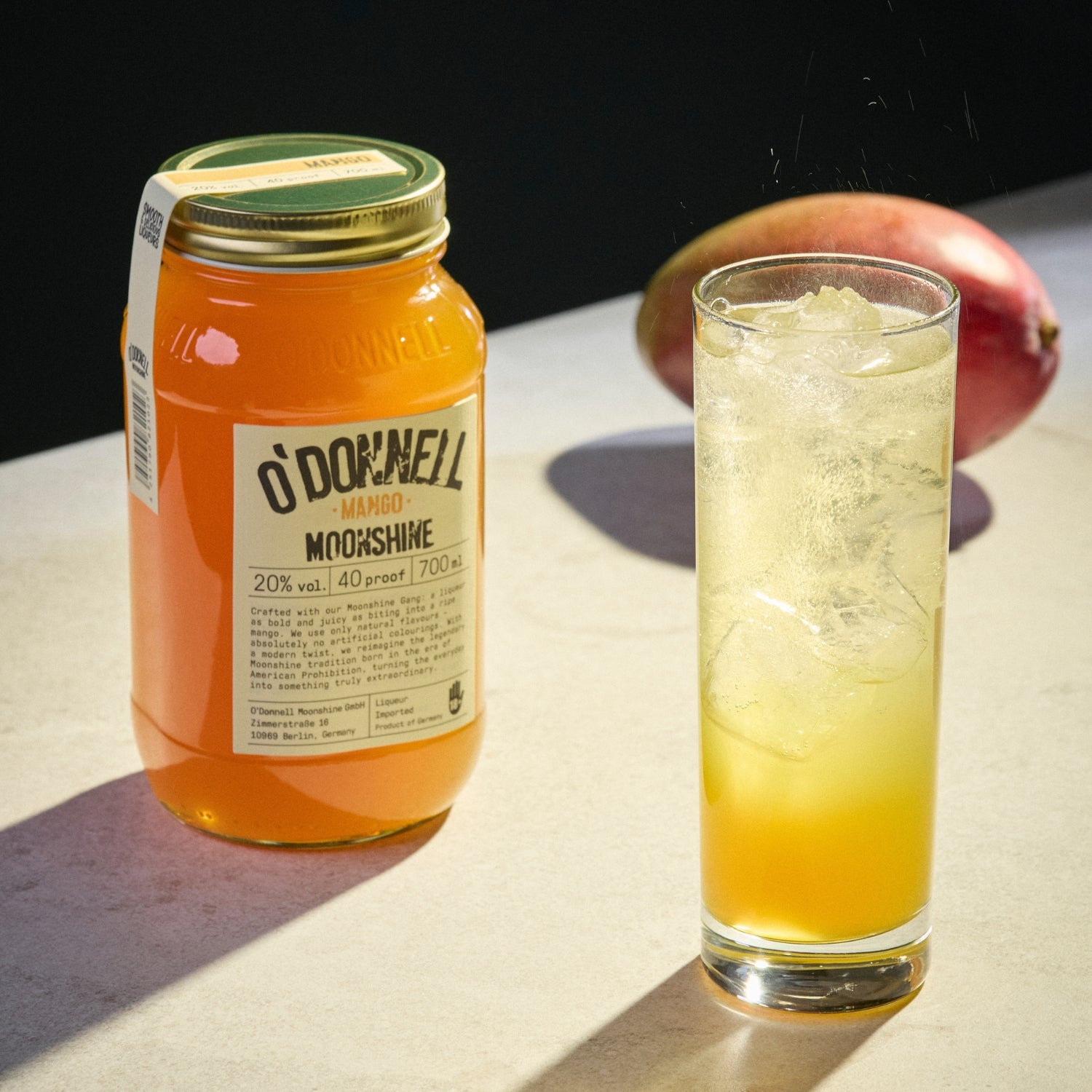 A Mango Liqueur on the left side and a glass filled with ice and the liqueur on the right side with a real Mango blurred in the background.