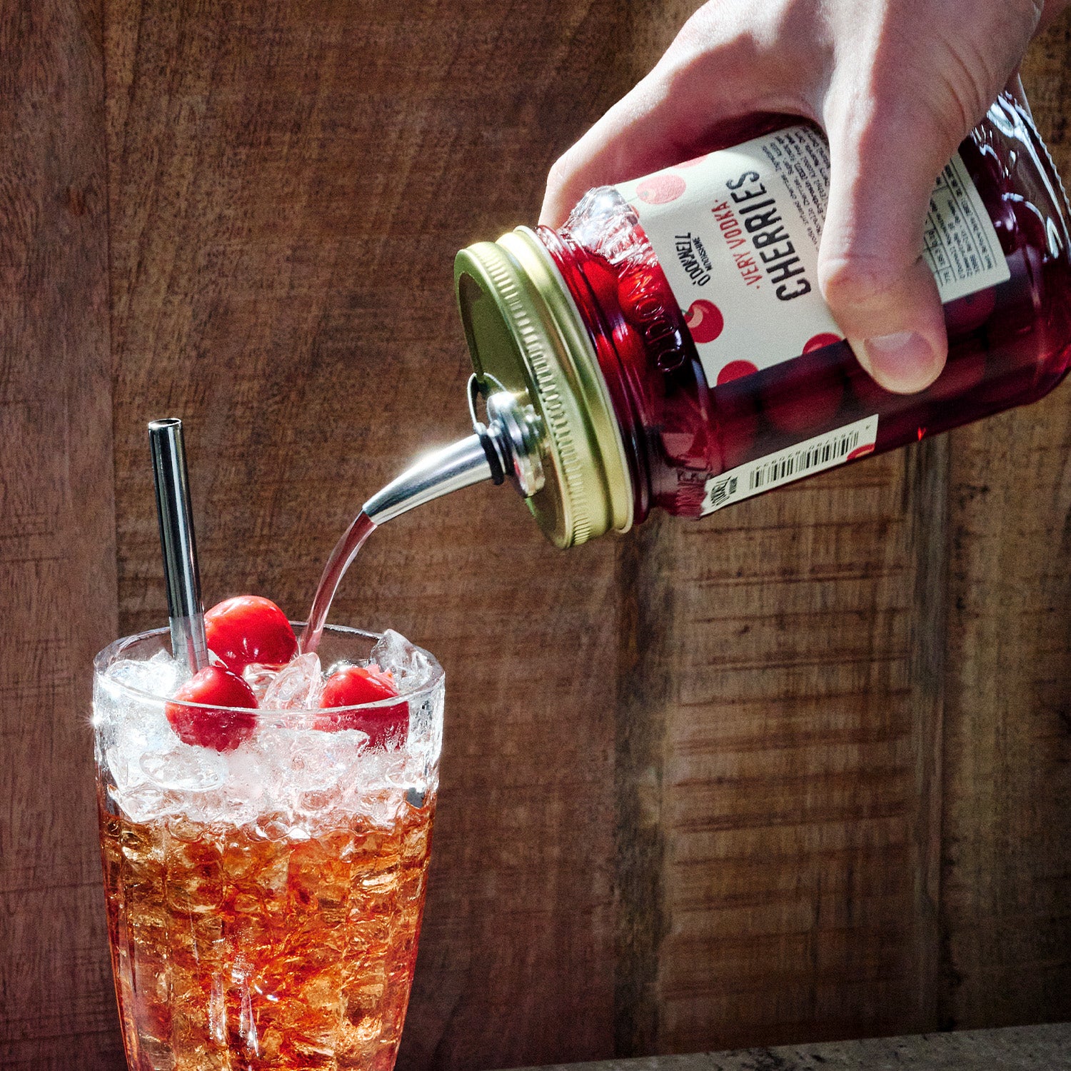 Hand pouring Very Vodka Cherries into a glass of iced drink with cherries on a wooden background