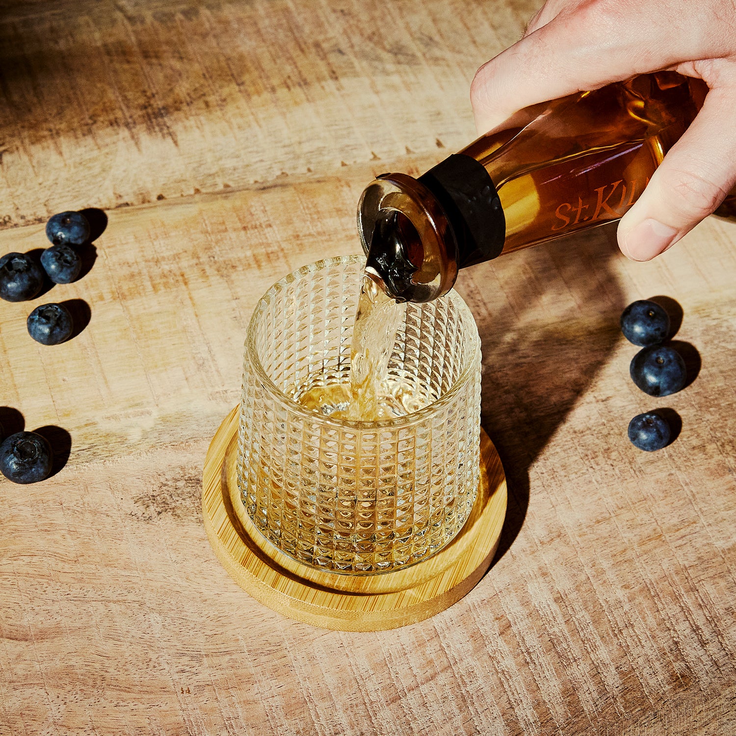 Textured Tumbler Glass on a wooden coaster. Surrounded by blueberries having liquid poured into it