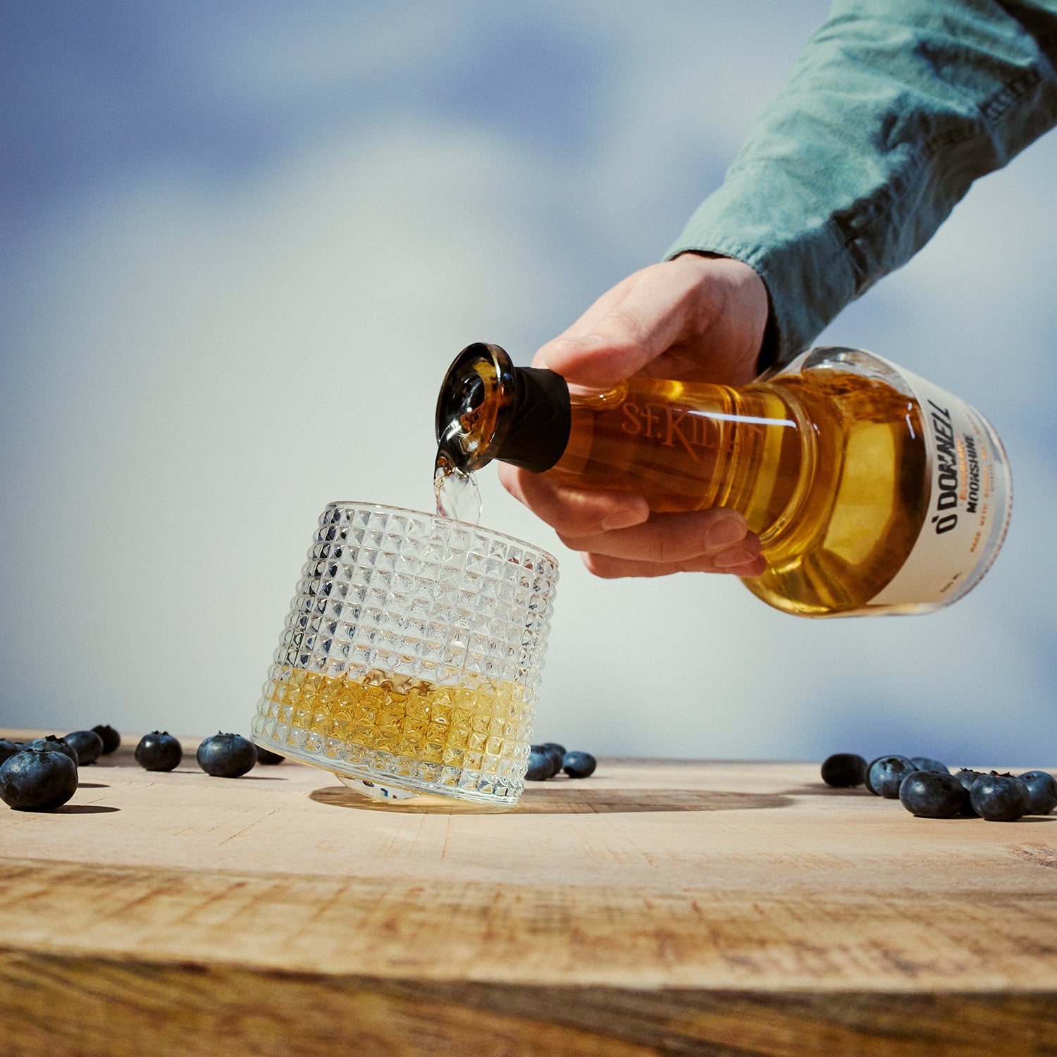 Hand pouring whiskey from a bottle into a textured tumbler glass on a wooden surface with blueberries.