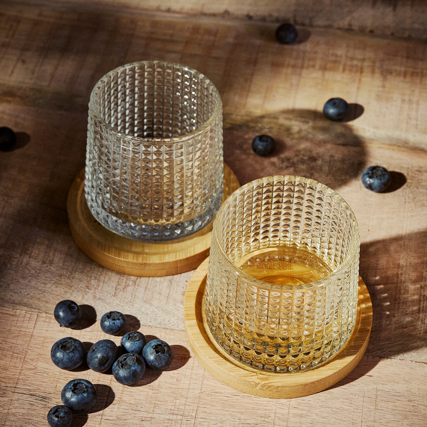 Two textured Tumbler Glasses on a wooden coaster. Surrounded by blueberries