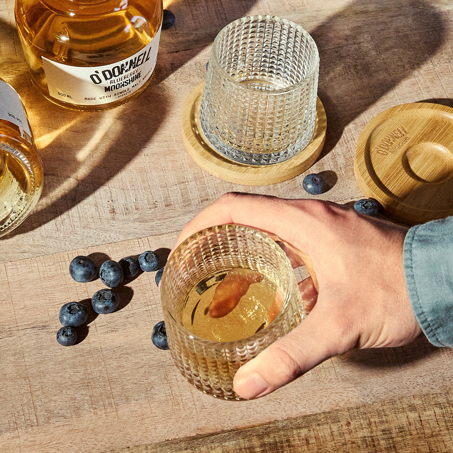 Person holding a textured tumbler glass of whiskey with blueberries on a wooden table, featuring O'Donnell whiskey bottle.