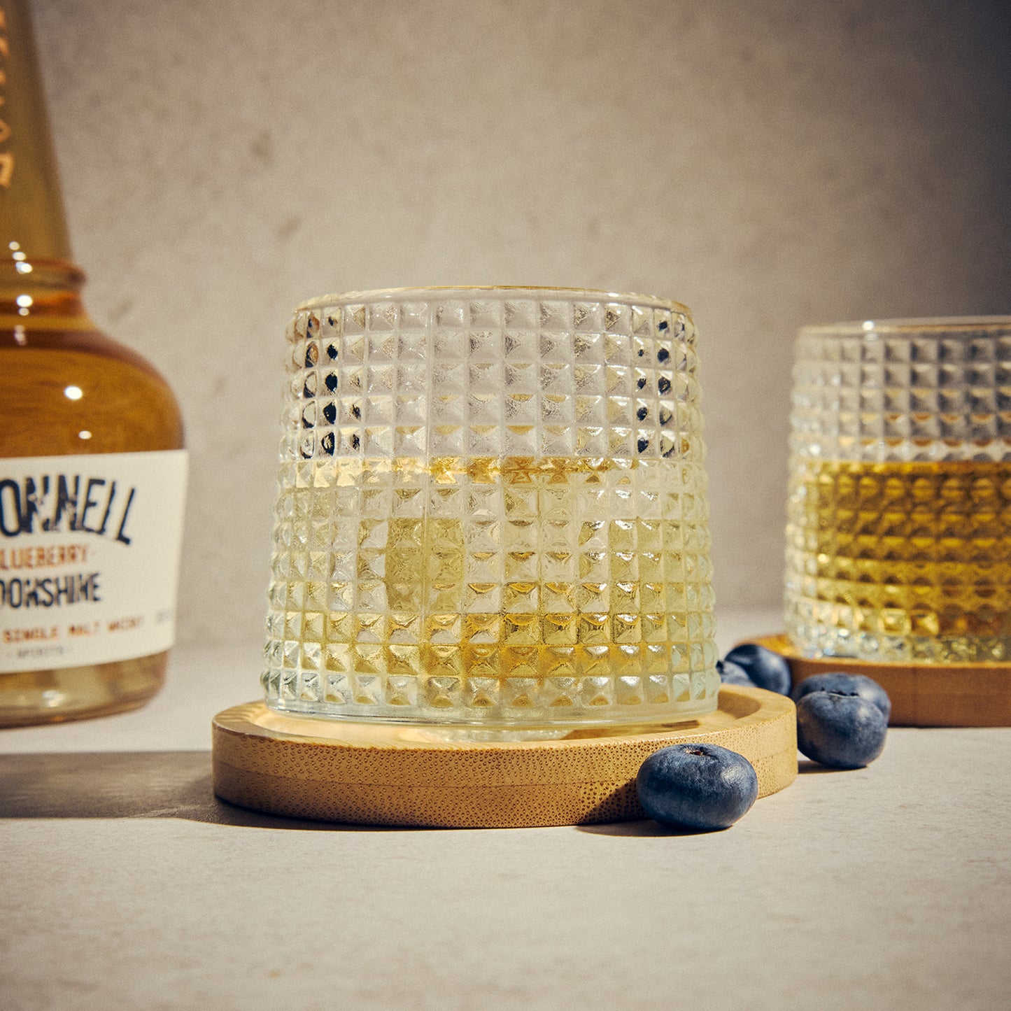 Two Textured Tumbler Glasses on a wooden coaster. Surrounded by blueberries