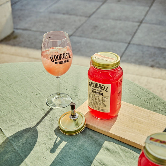 A blood Orange moonshine 700l Jar next to a O'Donnell Moonshine Spritz glass and a pouring lid.