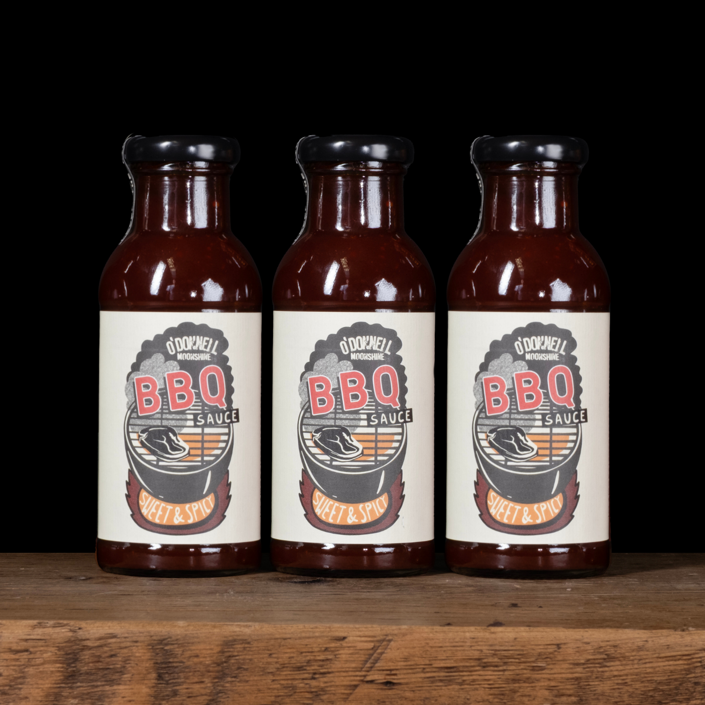 Three bottles of O'Donnell BBQ Sauce on a wooden surface with a black background