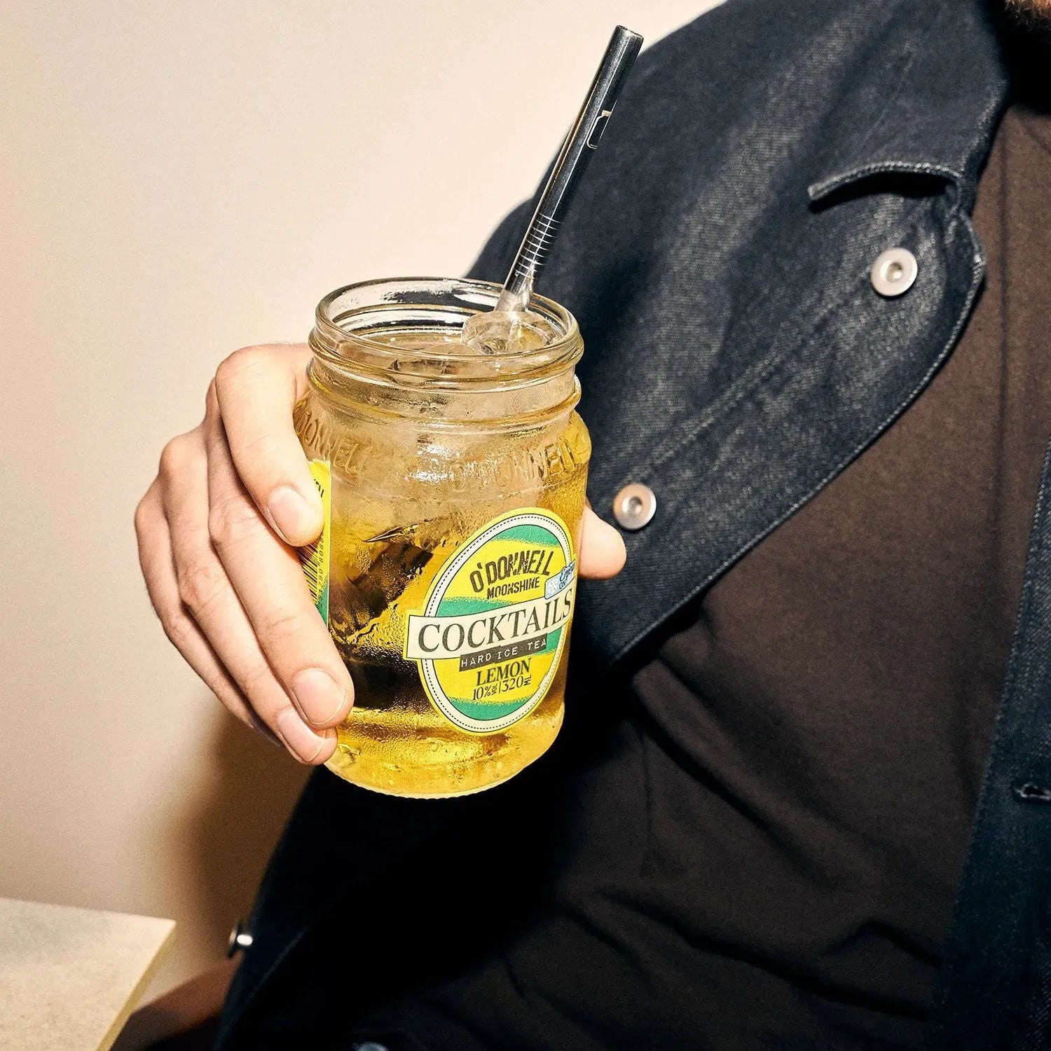 Person holding a jar of cocktails with a straw, wearing a denim jacket.